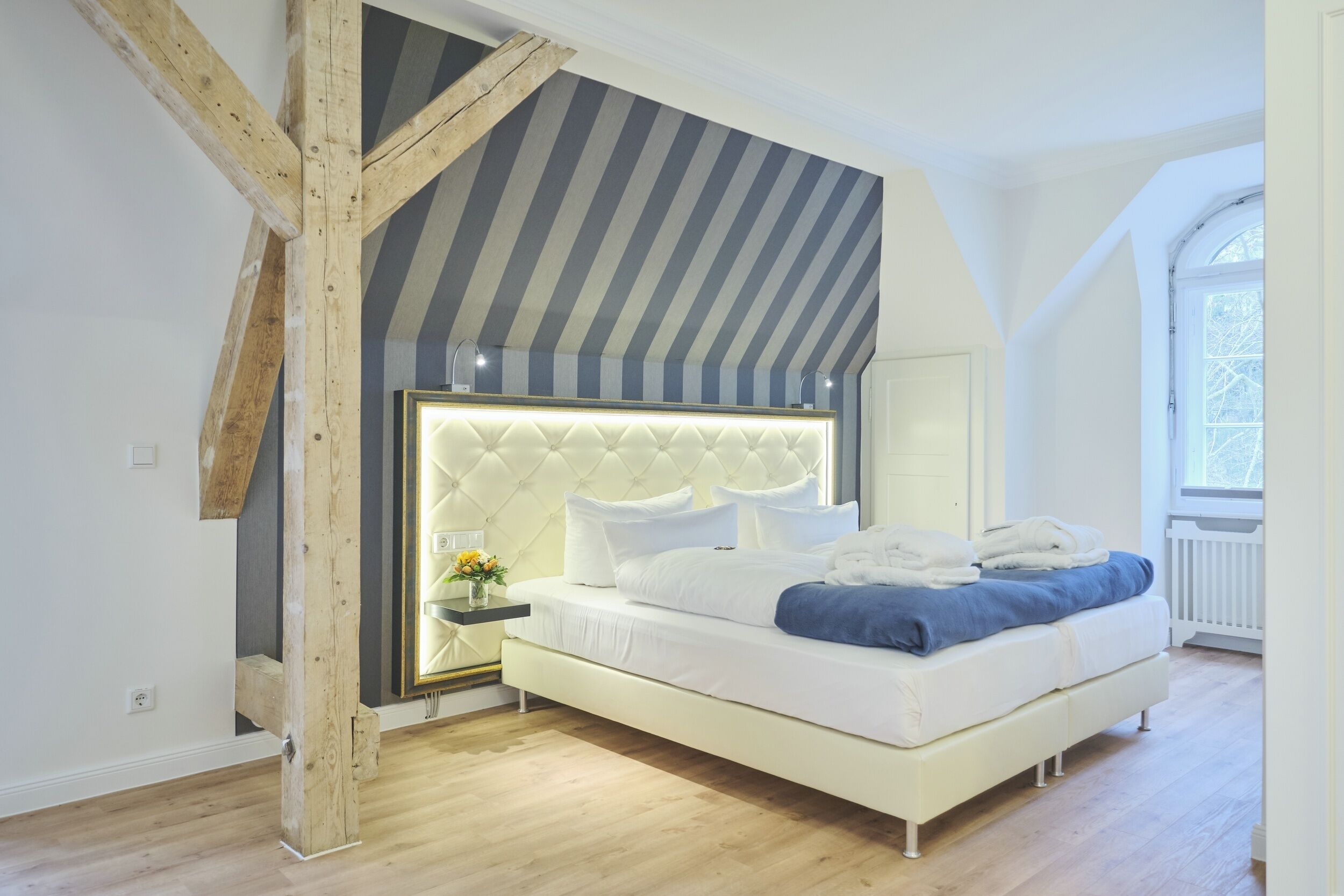 Un dormitorio moderno con techo inclinado, cabecera capitonada, cama blanca y vista al exterior a través de una ventana.
