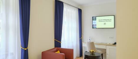 A hotel room with a bed, a red armchair, a small table with a lamp, a desk, and a TV displaying a welcome message.