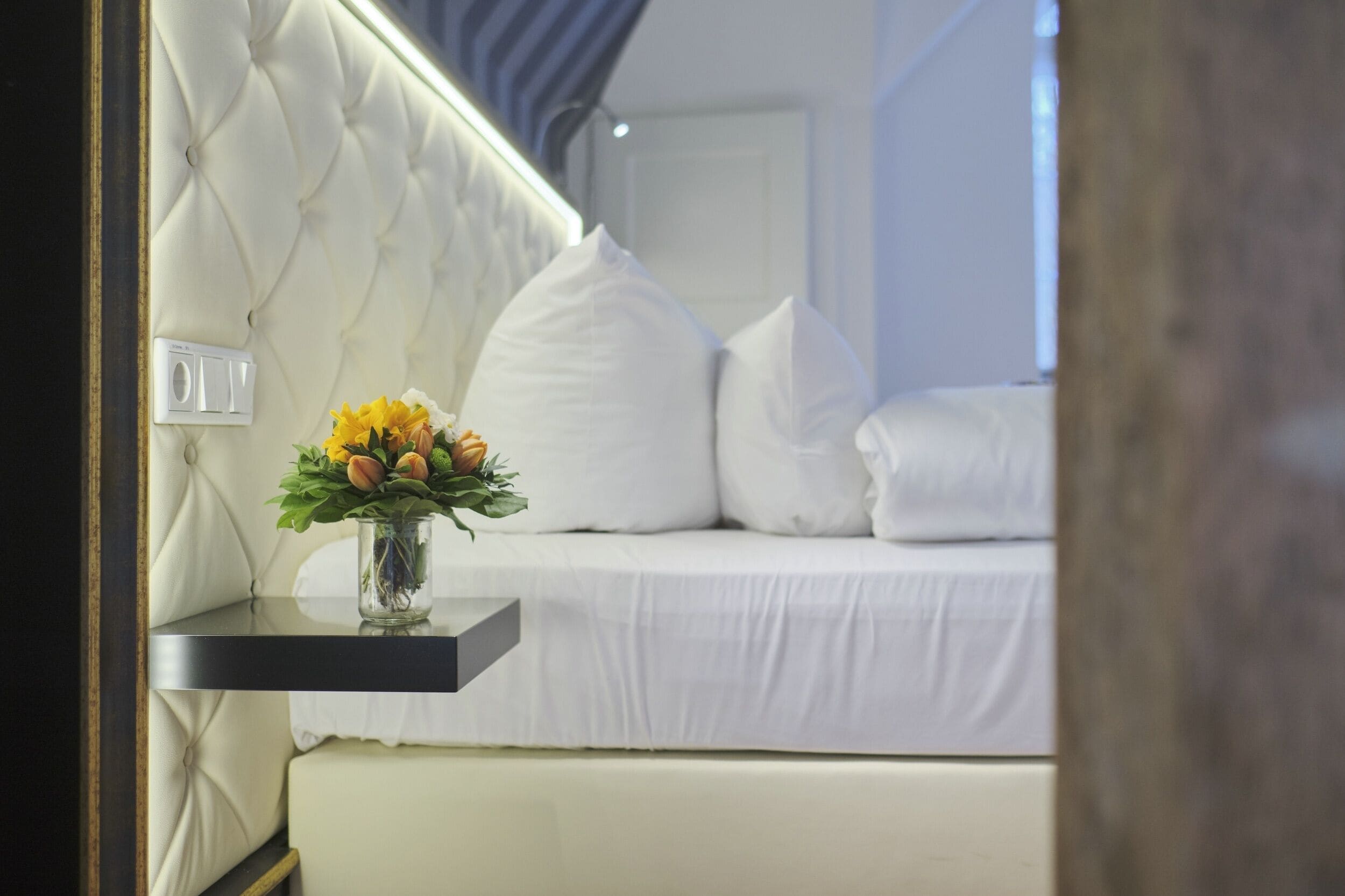 Un dormitorio con cabecera capitonada, una mesita de noche con un florero y una cama con ropa de cama blanca.