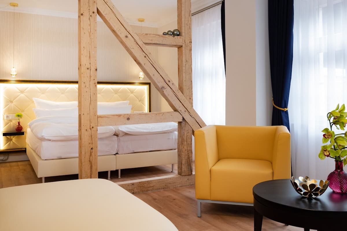 A hotel room with a bed, a yellow armchair, a small table with a vase, and a wooden staircase.