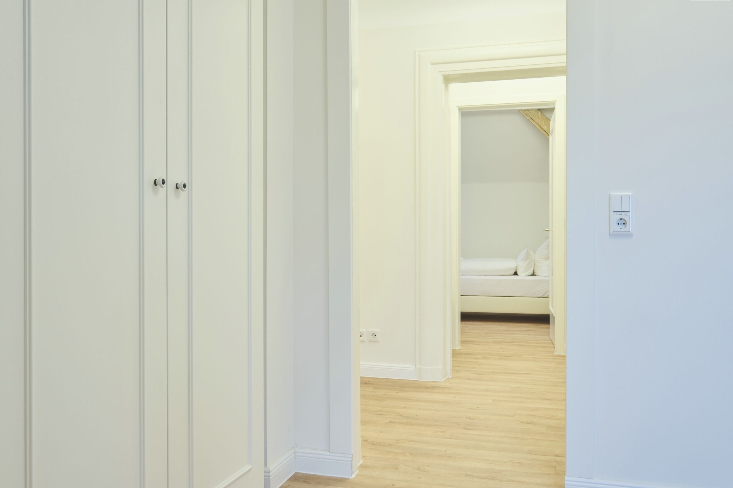 A hallway with white walls and wooden flooring leading to a bedroom with a bed and a nightstand.