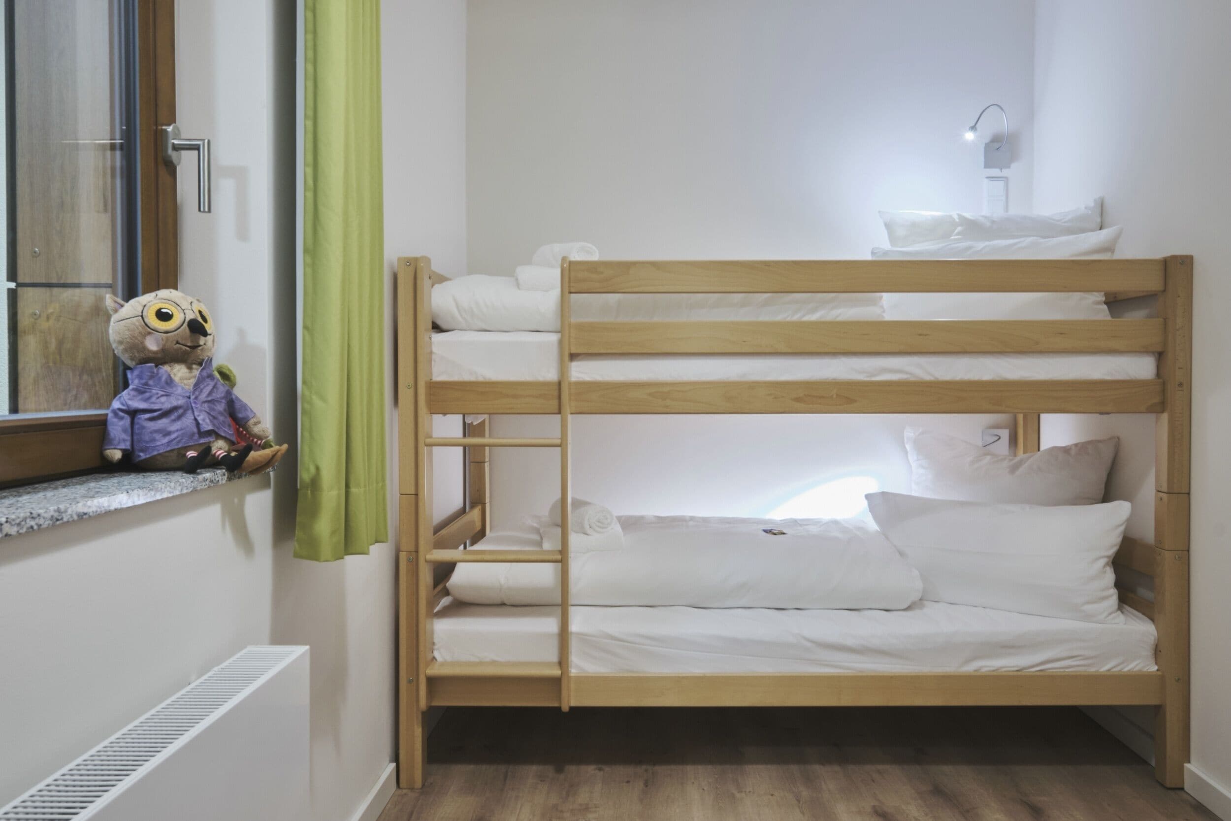 A bunk bed with white bedding, a window with a green curtain, and a teddy bear on a ledge.