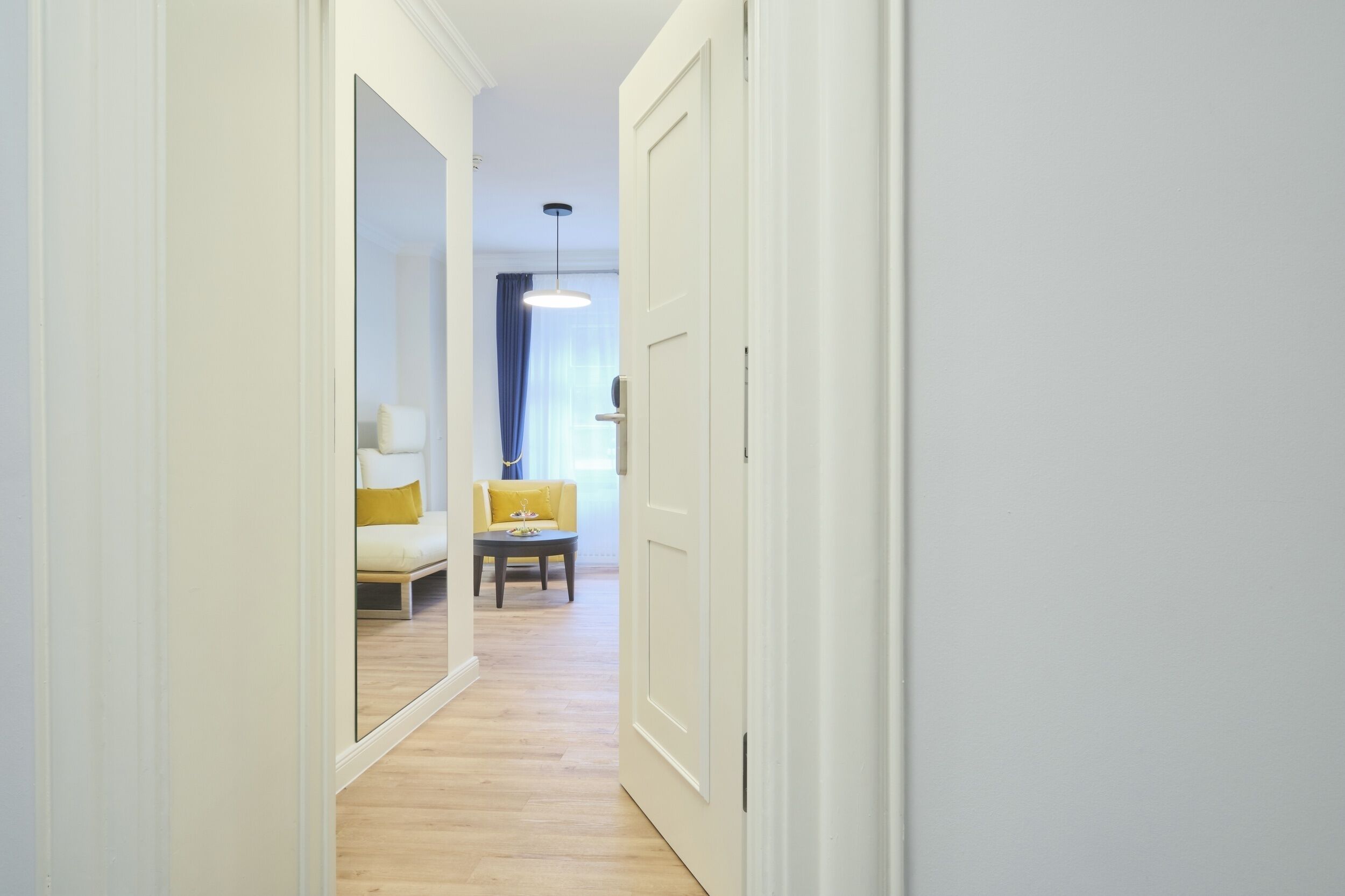 A hallway with a wooden floor, white walls, and a yellow sofa in the background.