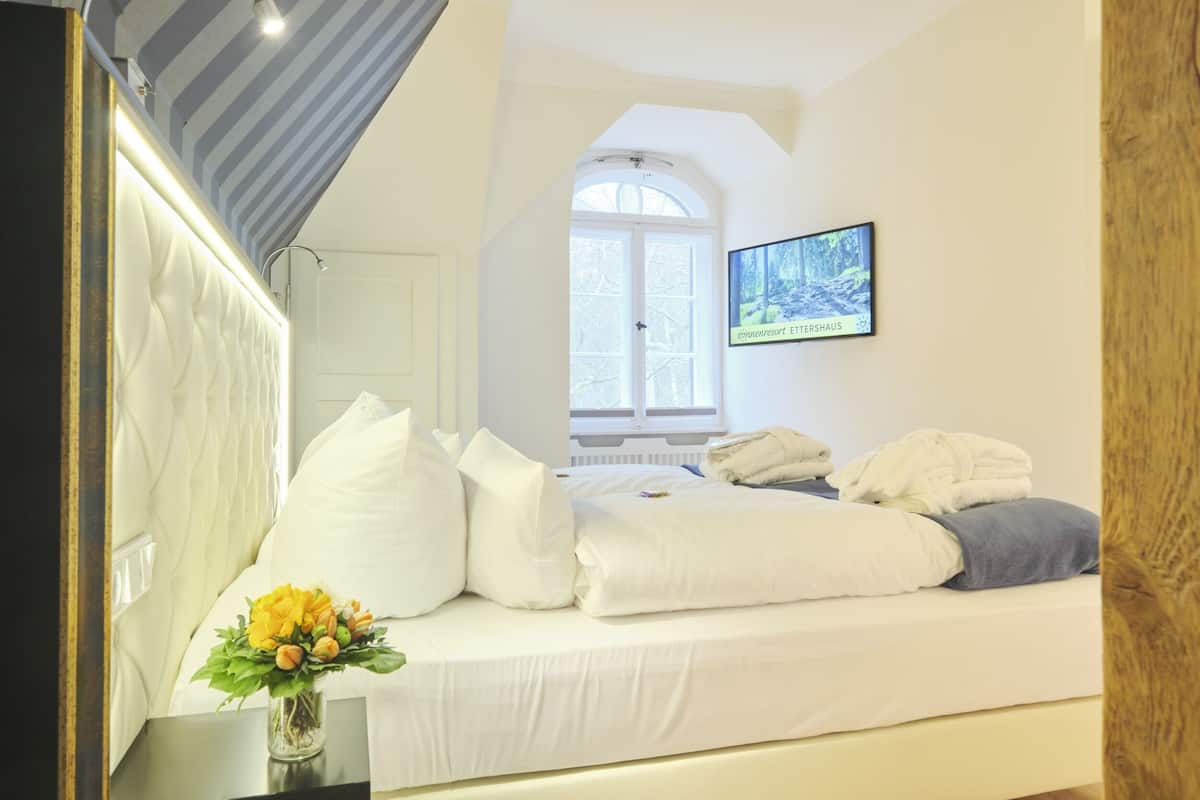 A neatly made bed with white linens and pillows, a small vase with yellow flowers, and a television mounted on the wall.