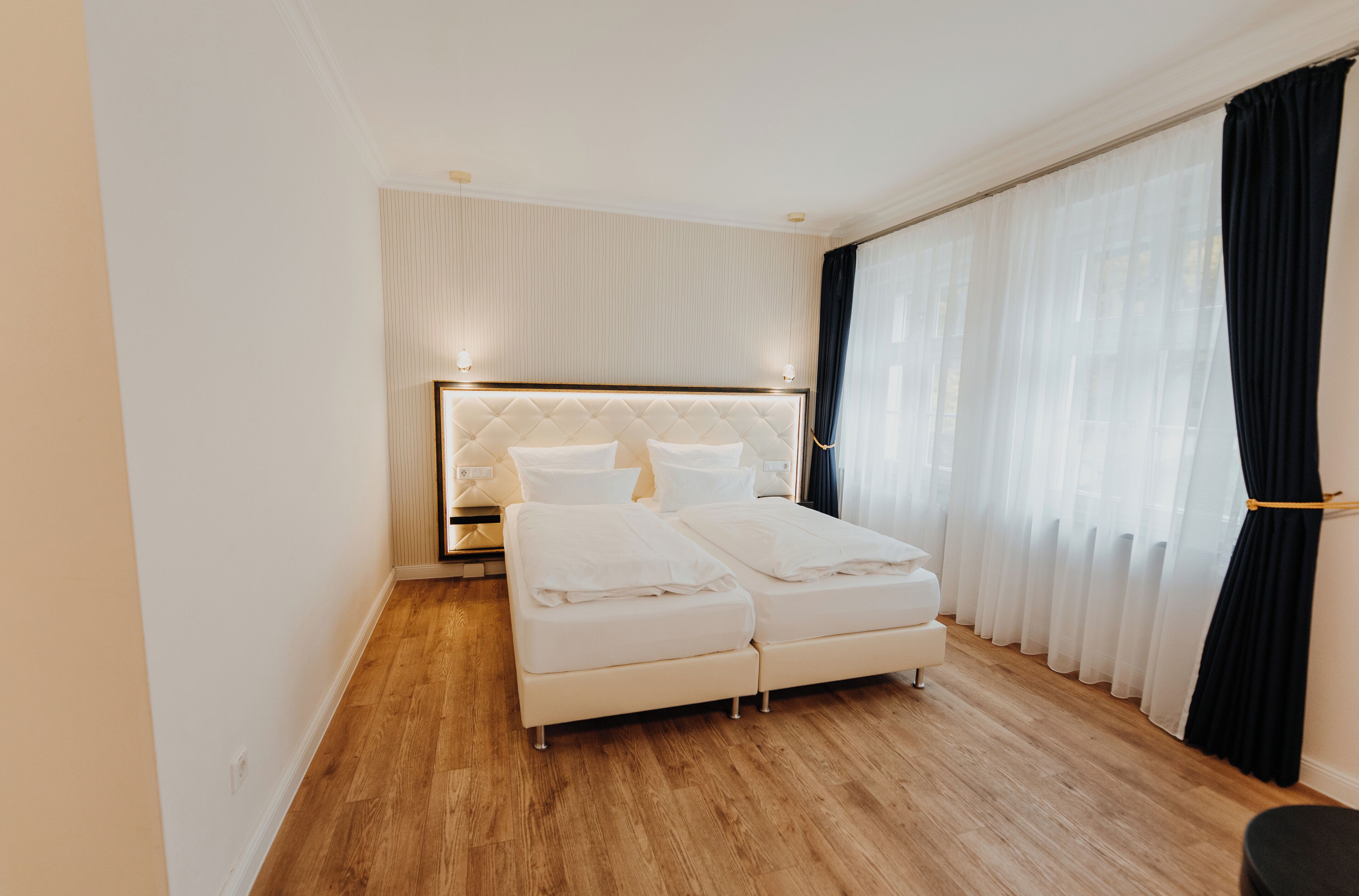 A modern hotel room with a large bed, a nightstand, and a window with sheer curtains.