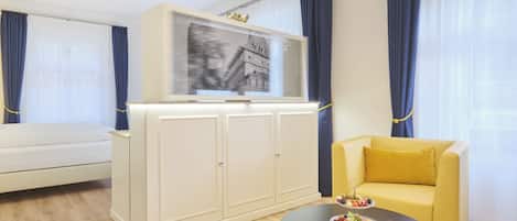 Una sala moderna con un sofá, una mesa de comedor con fruta, un televisor sobre un mueble bajo y un ventanal con cortinas.