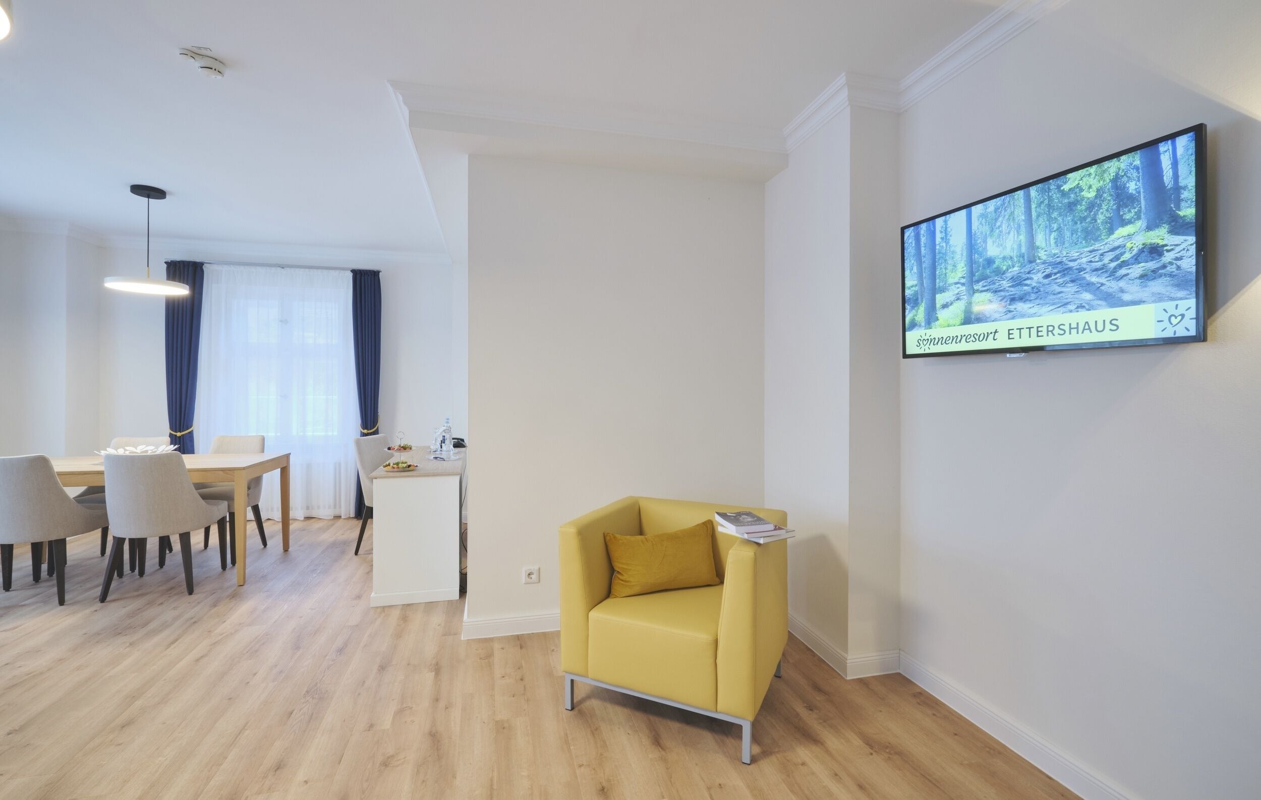 A modern living room with a yellow armchair, a dining area with a table and chairs, and a flat-screen TV on the wall displaying a forest scene.
