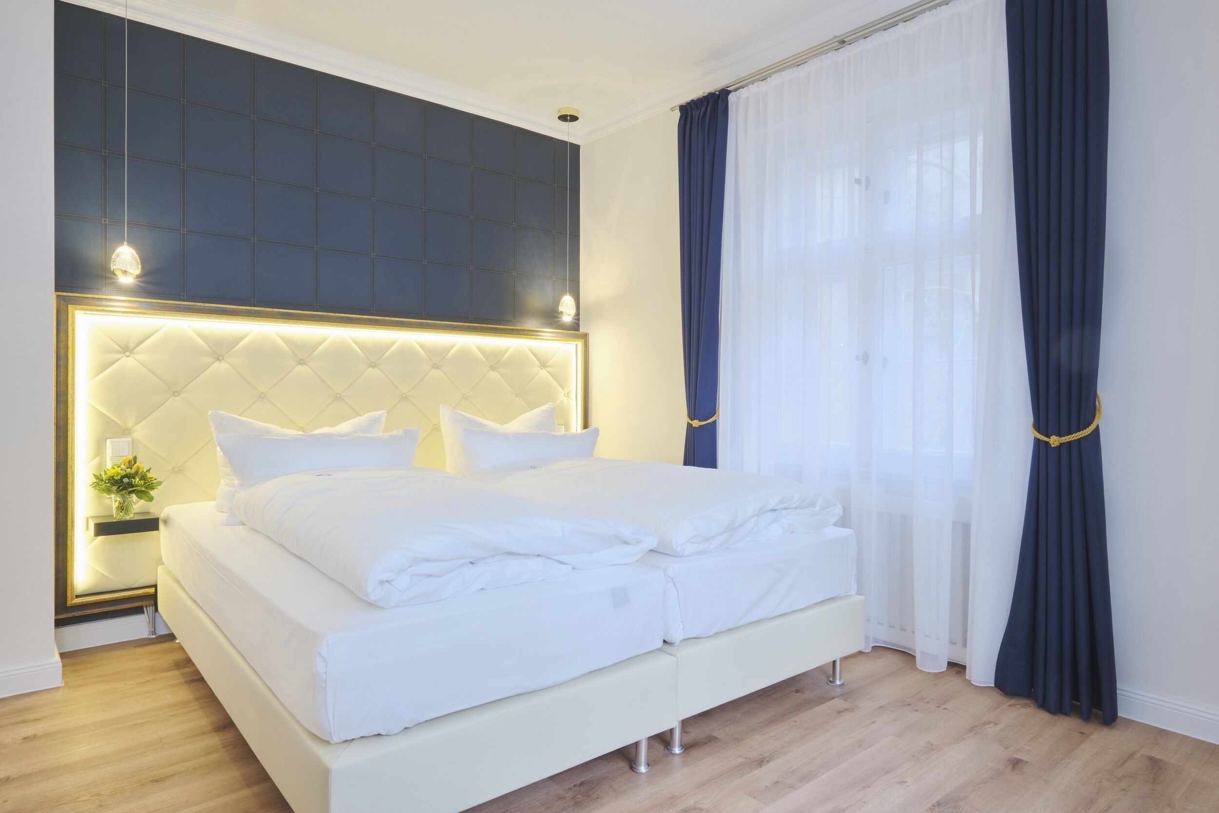 A neatly made bed with a tufted headboard, a bedside table with a lamp, and a window with sheer curtains.