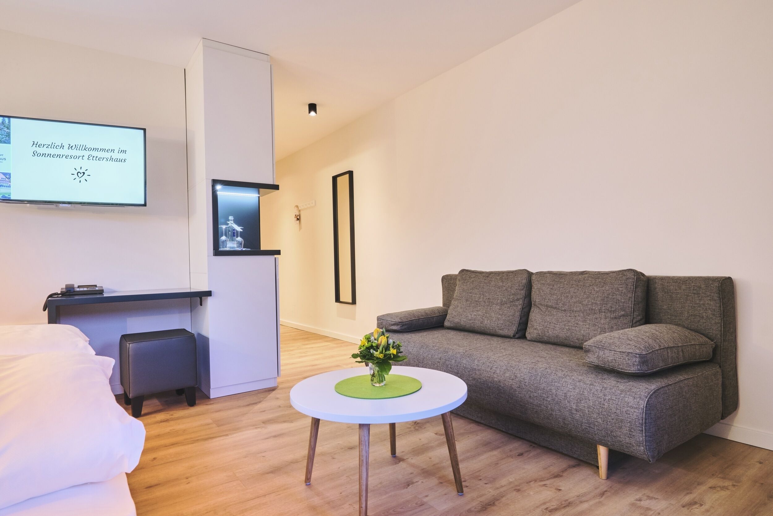 A modern hotel room with a grey sofa, a small round table with a green coaster, a bed with white bedding, a desk with a computer, and a TV displaying a welcome message.