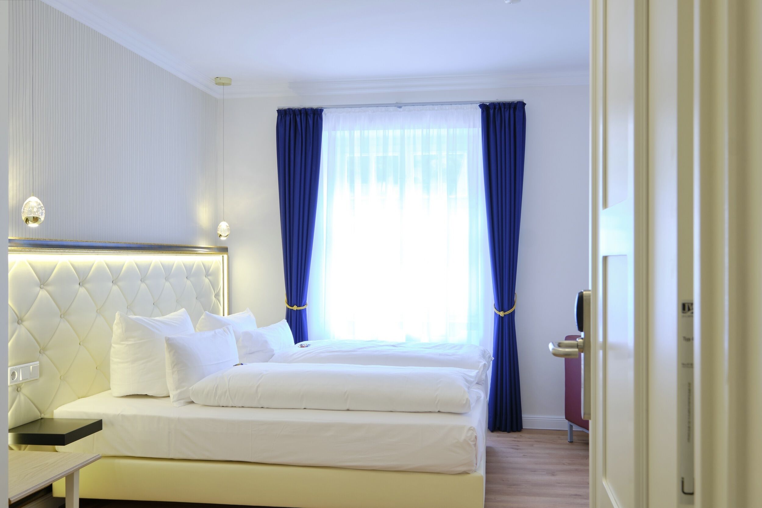 A hotel room with a large bed, a bedside table, and a window with blue curtains.