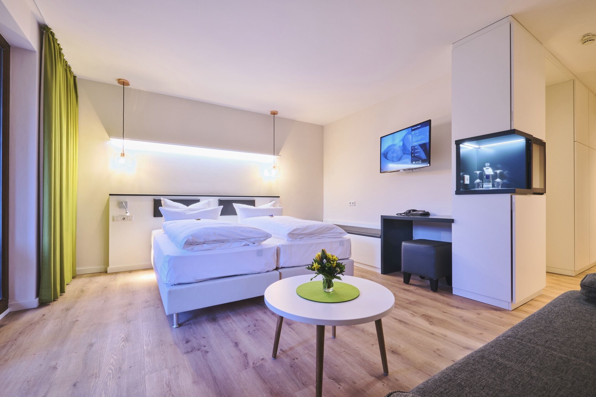 A modern hotel room with a bed, a desk, a TV, and a small table with a vase.
