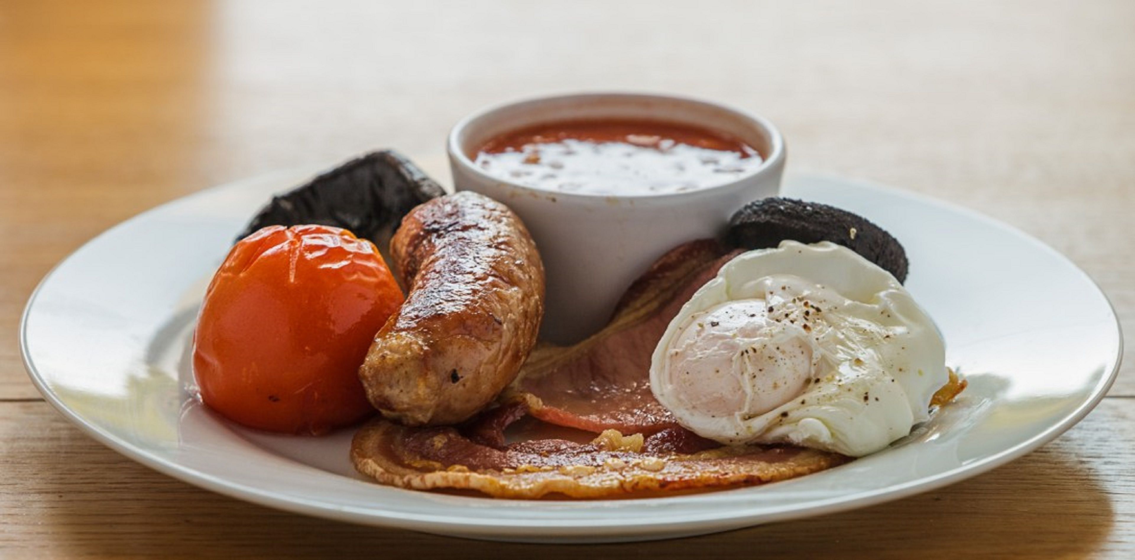 Un plato con una salchicha, un huevo pochado, tocino y tomate.
