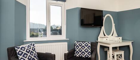 A room with a window, a television, a desk with a mirror, and two chairs with patterned cushions.