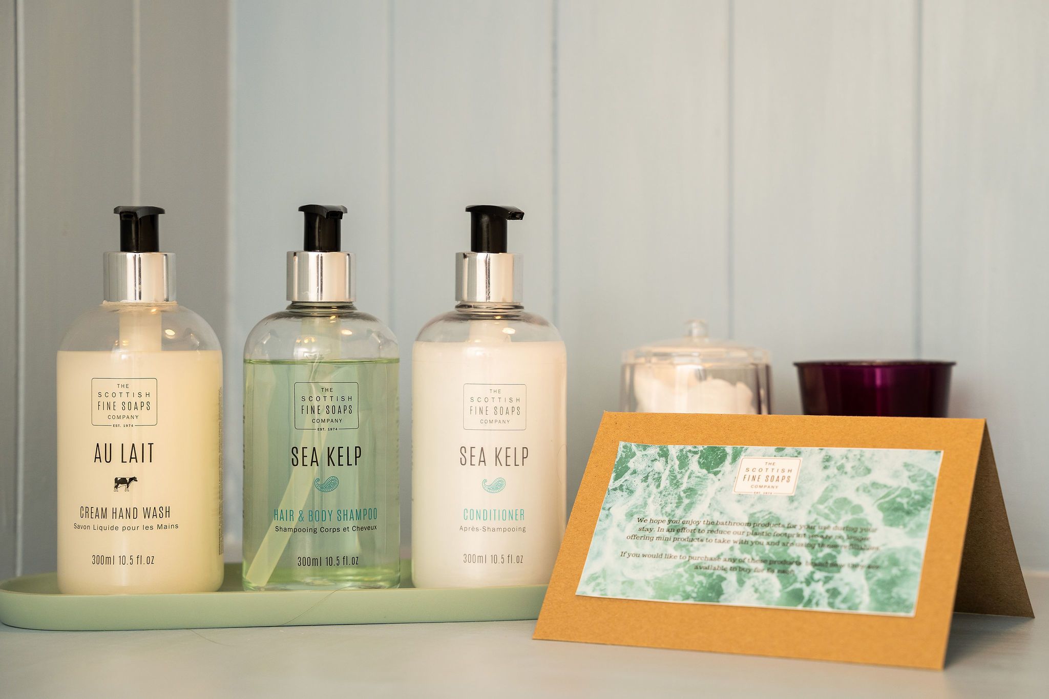 Tres productos de The Scottish Fine Soaps Company: jabón líquido para manos AU Lait Cream, champú y acondicionador de Algas Marinas, junto con una tarjeta y una vela.