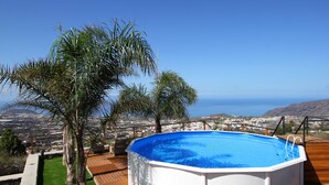Une piscine extérieure dotée d’une terrasse en bois, entourée de palmiers et offrant une vue sur une ville côtière.