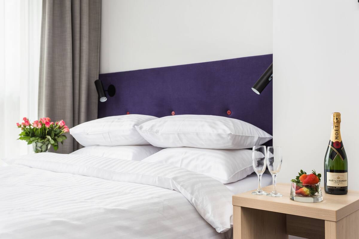 Un lit bien fait, avec du linge de lit blanc, une tête de lit violette et une table de chevet sur laquelle se trouvent une bouteille de champagne, un verre et un panier de fruits.