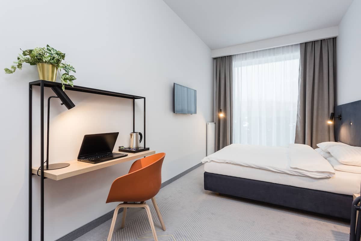 Une chambre d’hôtel avec un lit, un bureau avec un ordinateur portable, une chaise, une plante en pot et un téléviseur fixé au mur.