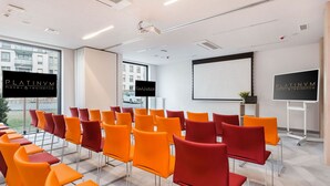 Una sala conferenze con file di sedie arancioni e rosse, uno schermo per proiettore e un cartello per Platinum Hotel & Residence.