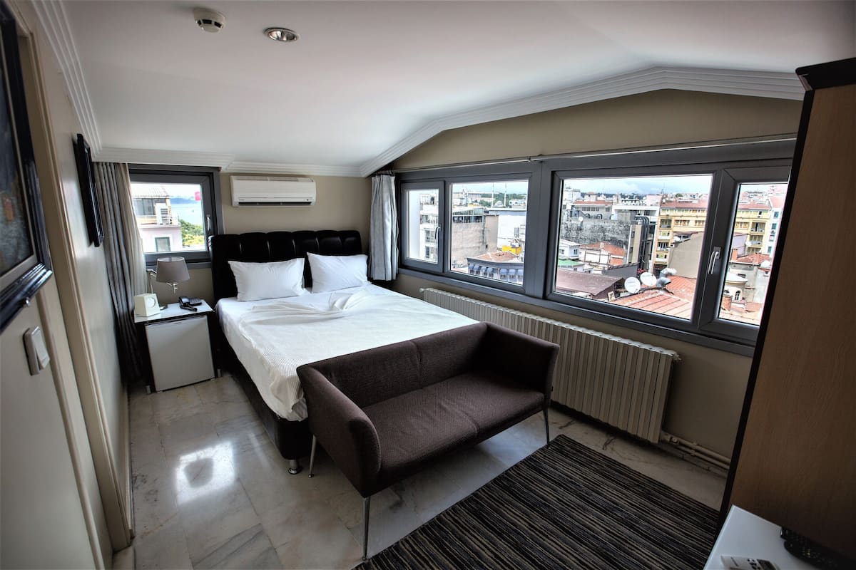 A hotel room with a bed, a small desk, a chair, a window with a city view, and a radiator.