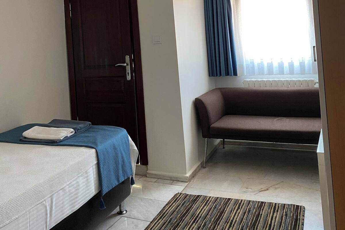 A hotel room with a bed, a sofa, a window with curtains, a radiator, and a door.