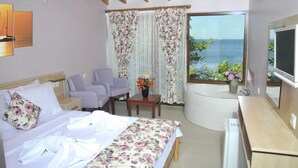 Habitación de hotel con cama, dos sillones, una mesita con un jarrón de flores, televisión y vista al mar a través de una ventana con cortinas.