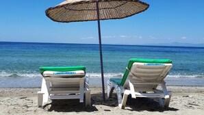 Dos sillas de playa con cojines verdes y una sombrilla de playa tejida sobre arena, con agua azul cristalina y un mar tranquilo al fondo.