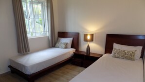 A hotel room with two single beds, a nightstand with a lamp, a window with curtains, and a wooden floor.