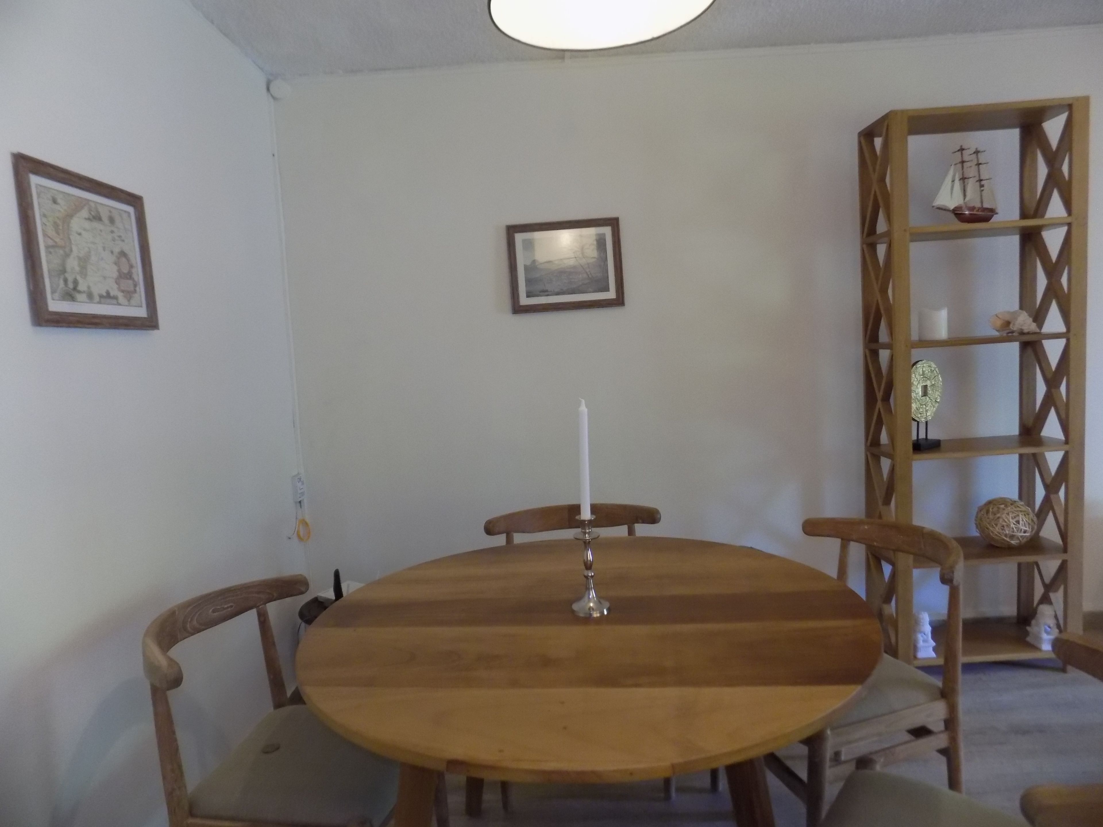 A round wooden dining table with four chairs, a white candle, and a small ship model on a wooden shelf.