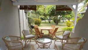 A covered patio with wicker furniture and a glass table, overlooking a garden with various plants and trees.