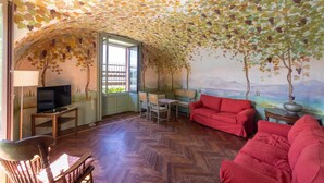 A cozy room with a herringbone floor, a red sofa, a television, and a mural of trees on the walls. - Villa La Rossa (Varenna)