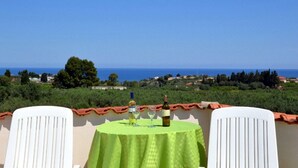 Une table recouverte d’une nappe verte, deux chaises blanches et une bouteille de vin, le tout sur une terrasse donnant sur un paysage côtier.