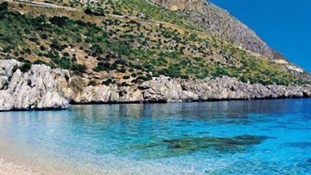 Une plage côtière bordée d’une eau d’un bleu limpide, d’une côte rocheuse et d’un paysage vallonné.