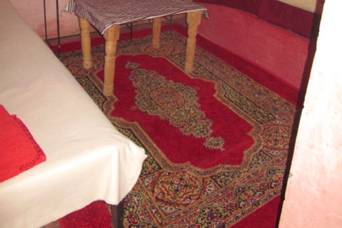 Une petite pièce, de taille modeste, comprenant un lit simple, une petite table, un banc, une fenêtre grillagée et un lavabo.