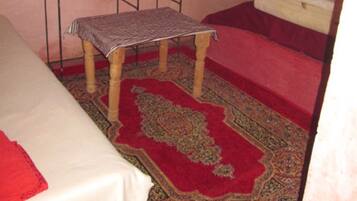 Habitación pequeña y compacta con una cama individual, una mesita, un banco, una ventana con barrotes y un lavabo.