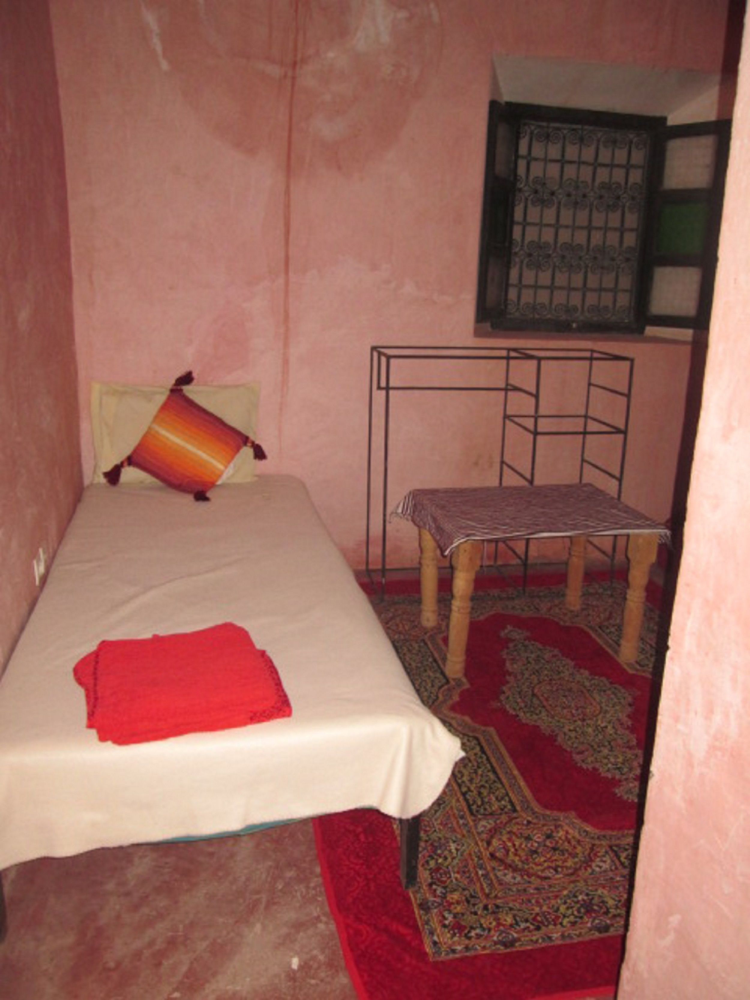 A small, single-bed room with a red blanket on the bed, a patterned rug on the floor, a metal chair, and a window with a metal grid.