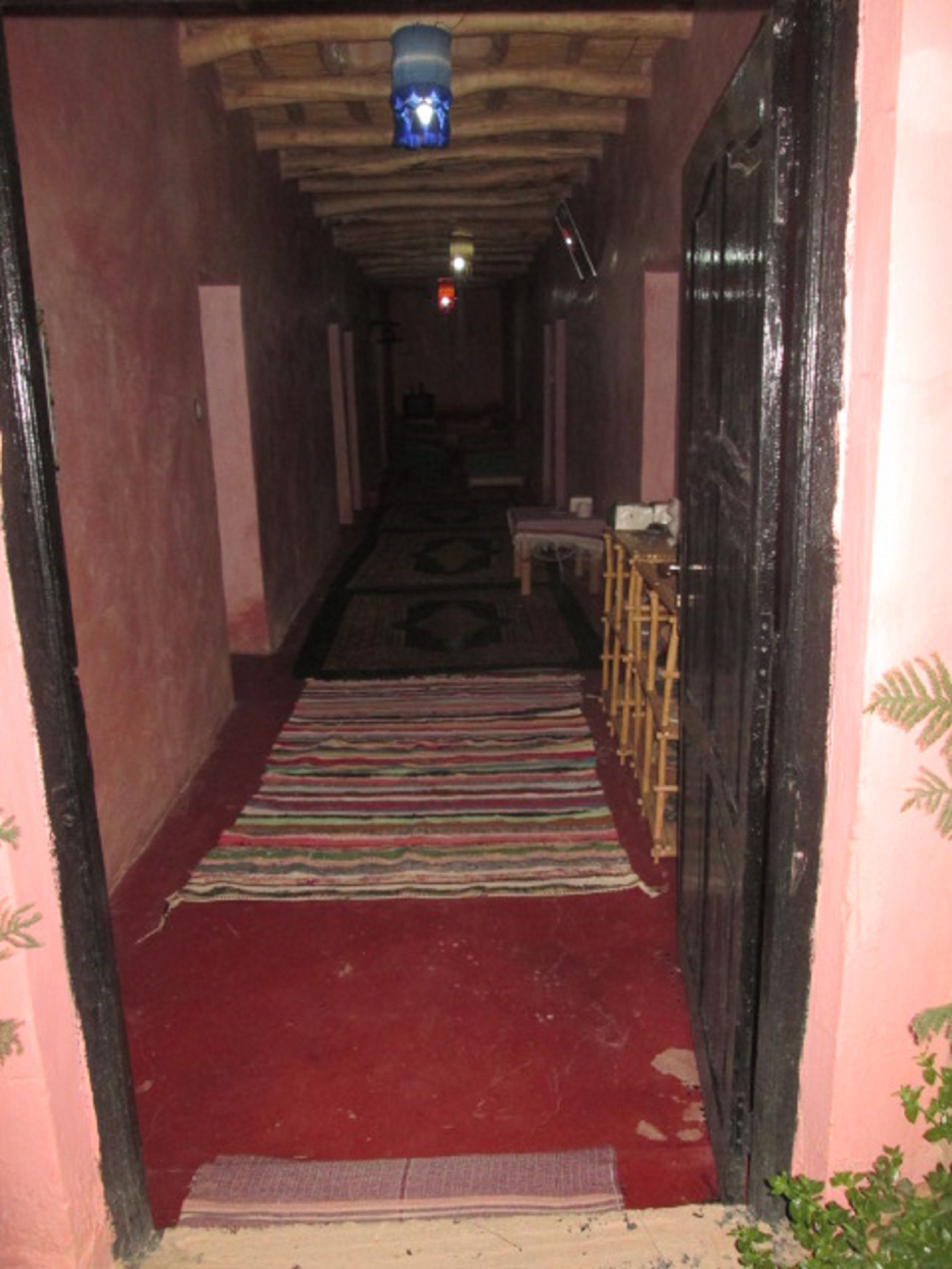 A narrow hallway with a red carpet leading to a door, wooden furniture, and a patterned rug.