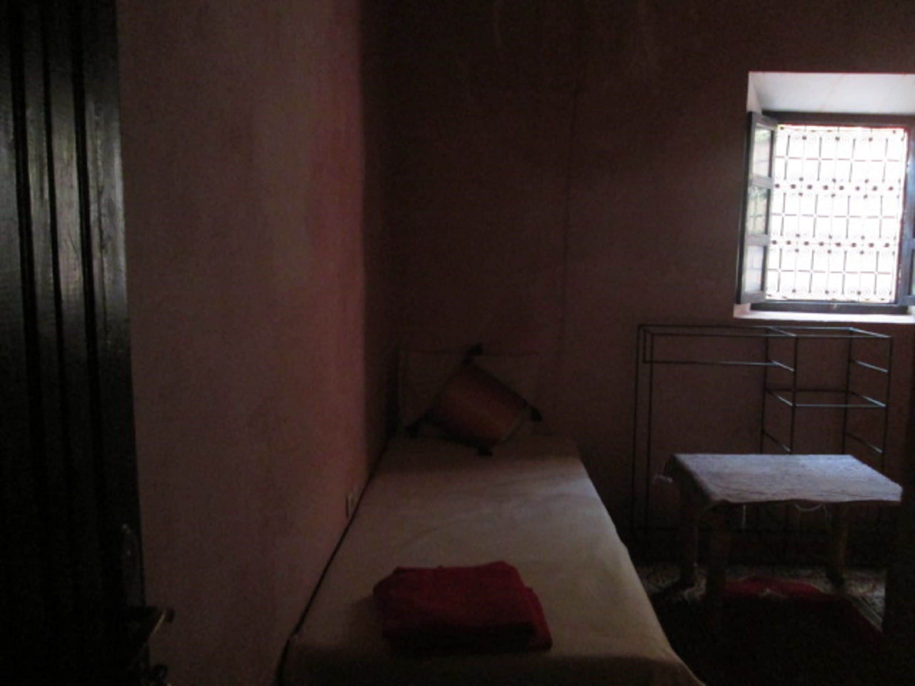 A narrow room with a single bed, a small table, and a window with a metal grid.