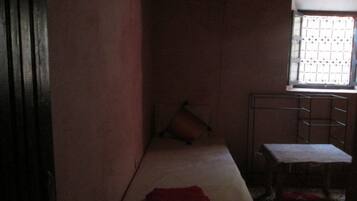 A narrow room with a single bed, a small table, and a window with a metal grid.