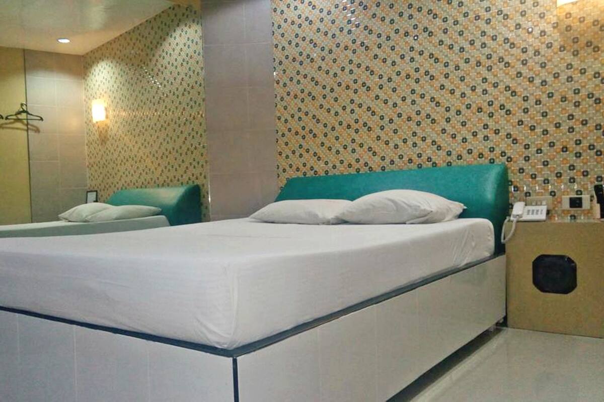 A hotel room with a bed, a bedside table, a mirror, and a wall with patterned wallpaper.