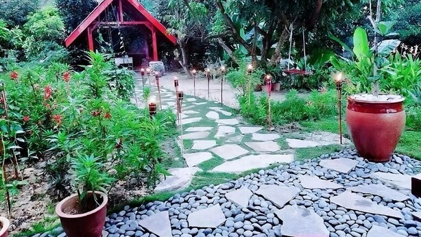 Un sendero en un jardín que conduce a una estructura de techo rojo, rodeada de abundante follaje y plantas en macetas.