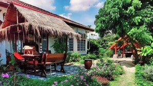 Una casa con techo de paja, un banco de madera y un jardín con flores rosadas y plantas verdes.