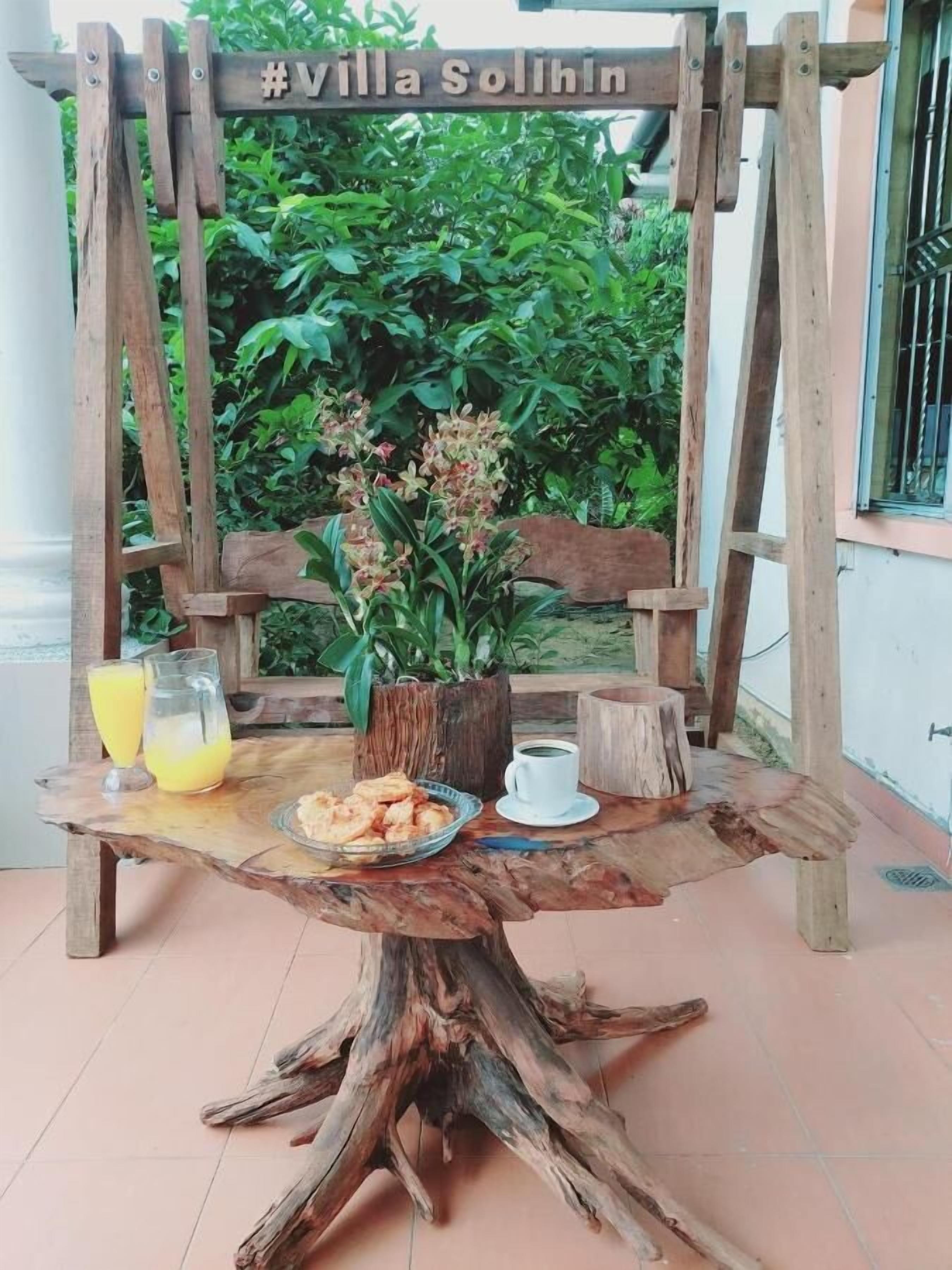 A rustic wooden table with a sign reading "#Villa Solihin" above it, surrounded by greenery.