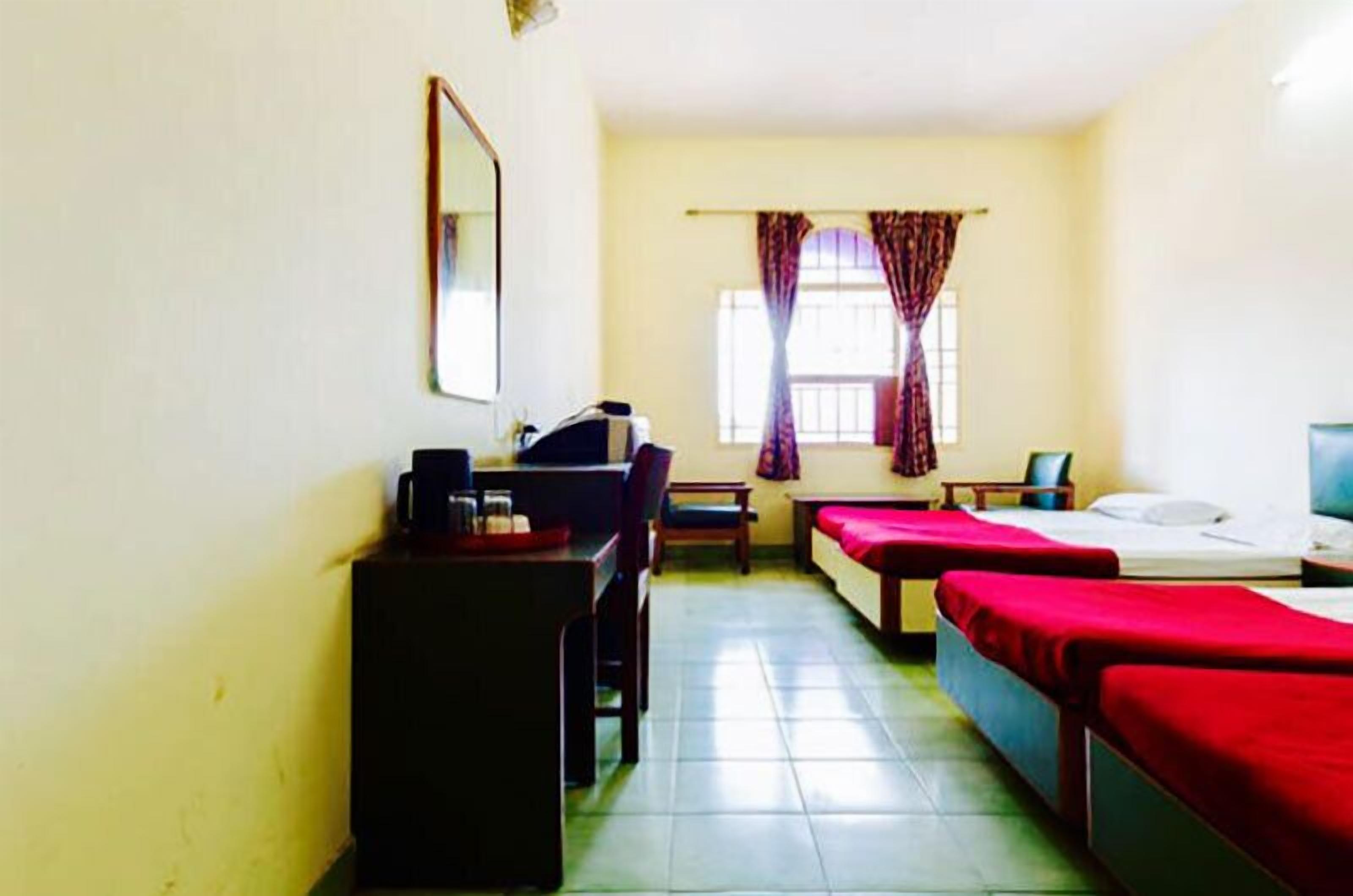 A hotel room with two single beds, a desk with a chair, a small table, and a window with curtains.