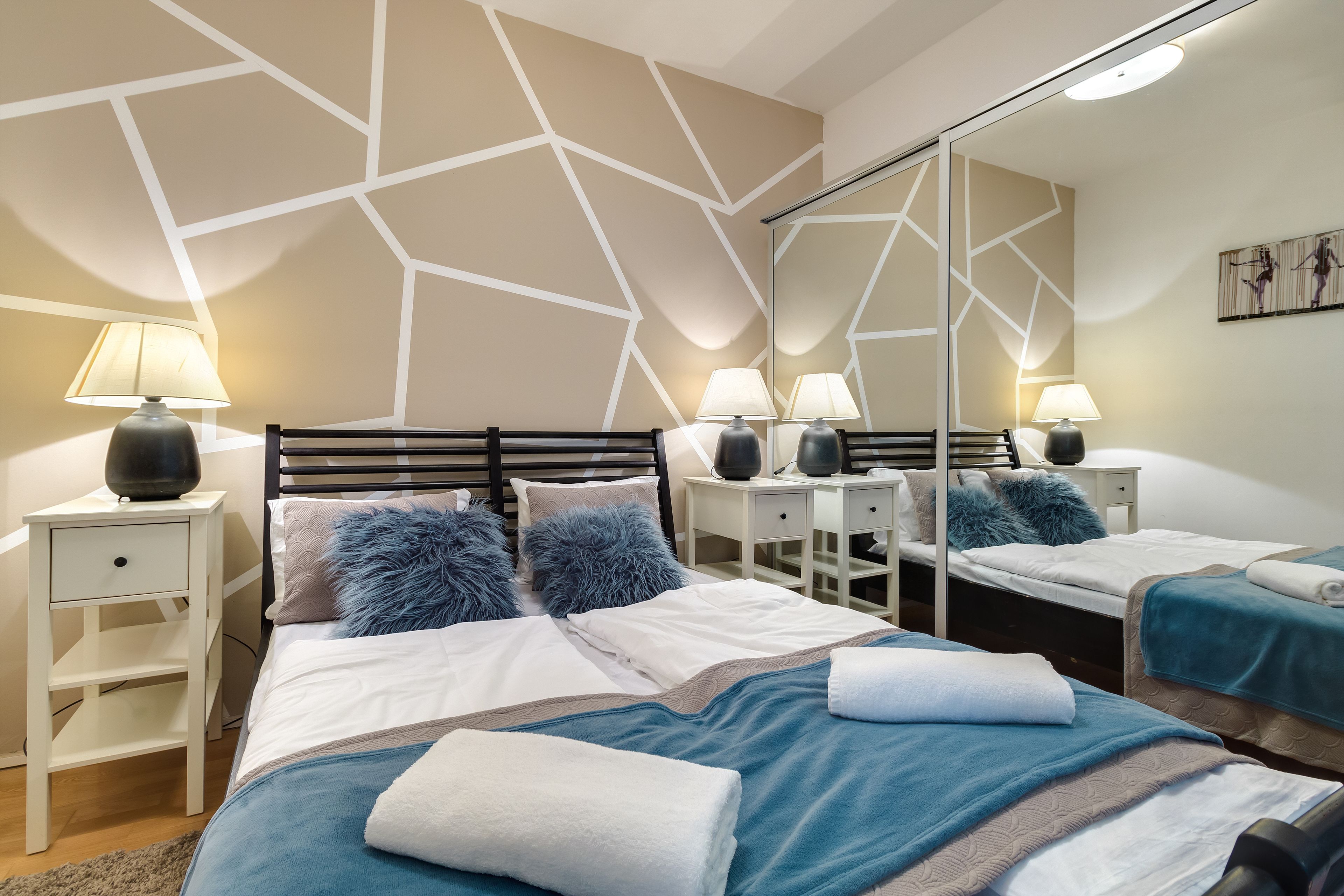 A modern hotel room with a large bed, two bedside tables with lamps, a mirror, and a geometric-patterned wall.