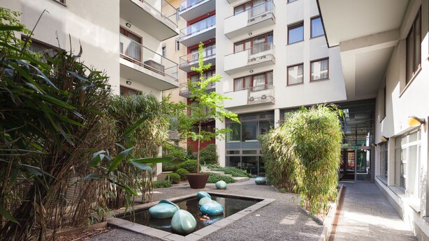 Un edificio moderno con varios balcones, un pequeño jardín con fuente de agua y un camino que lleva a la entrada.