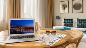 Un MacBook Air sobre una mesa redonda junto a un mapa y gafas, una lámpara y una cama con almohadas estampadas en una habitación con cortinas y obras de arte enmarcadas en la pared.