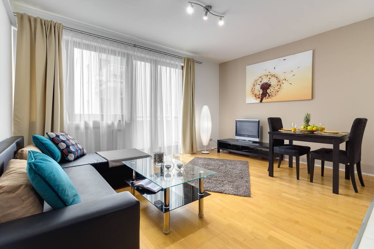 Una sala de estar moderna con una mesa de centro de vidrio, un sofá con cojines, un rincón de comedor con una mesa y sillas, un televisor de pantalla plana y un cuadro abstracto grande en la pared.