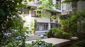 Un patio con un banco de hormigón, abundante vegetación y un edificio con varias ventanas y balcones.