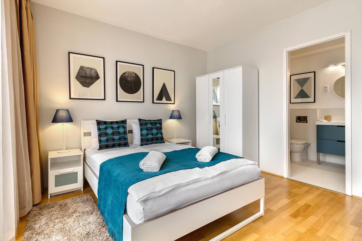 Un dormitorio moderno con cama, mesitas de noche, un armario y un baño visible a través de una puerta abierta.