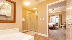 A bathroom with a bathtub, a glass shower enclosure, and a door leading to a bedroom with a bed and a chair.